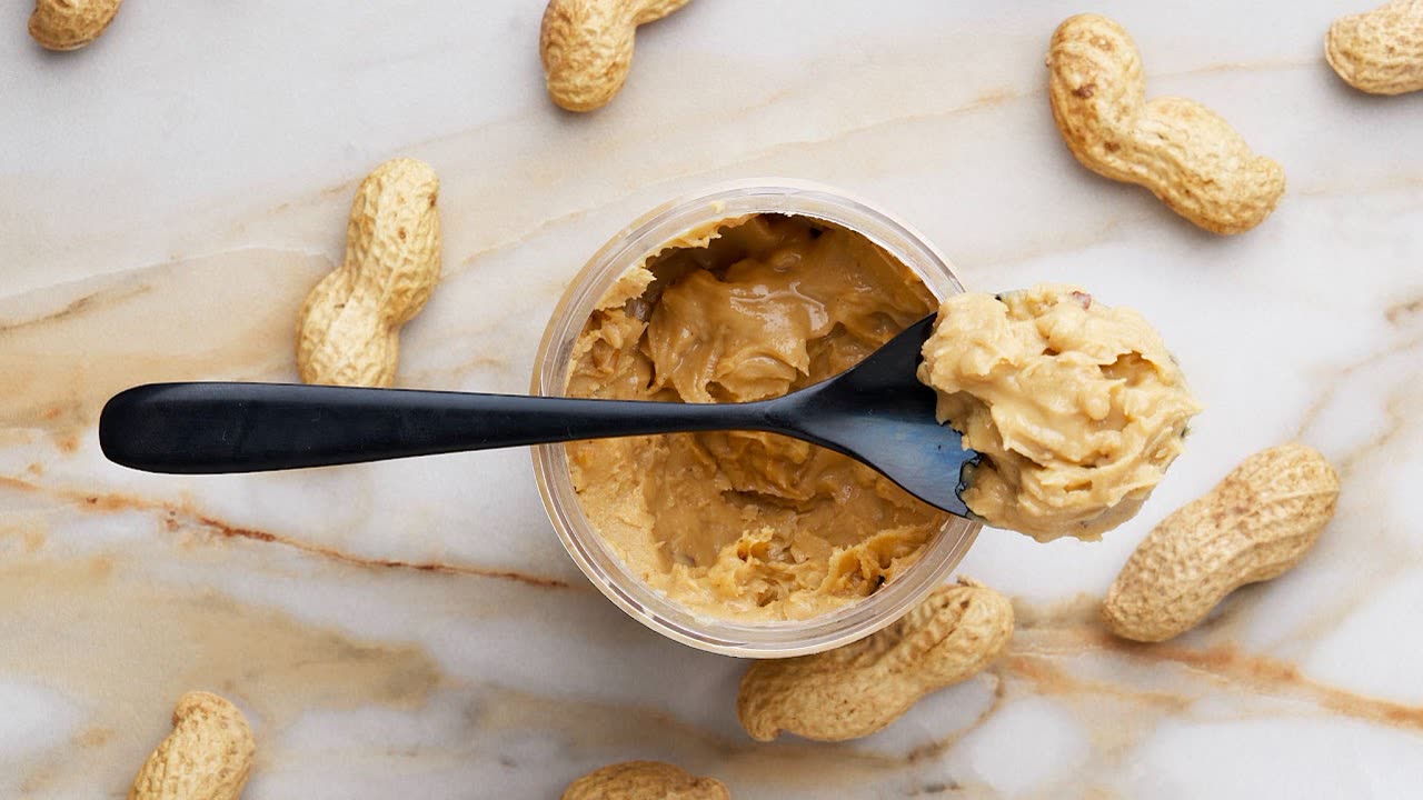 An open jar of peanut butter with a spoon, surrounded by in-shell peanuts